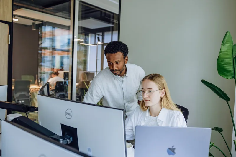 Two colleagues collaborating at a desk with computers in a modern office setting.