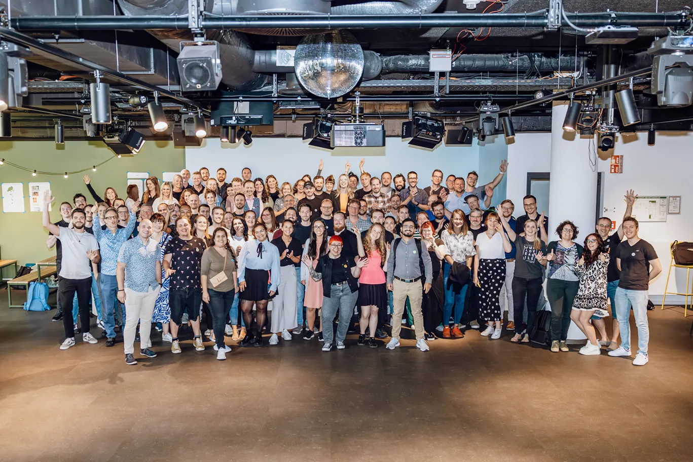 A large group of people posing together indoors, smiling and waving at the camera. The setting appears to be a modern office or event space with lighting and a disco ball overhead.