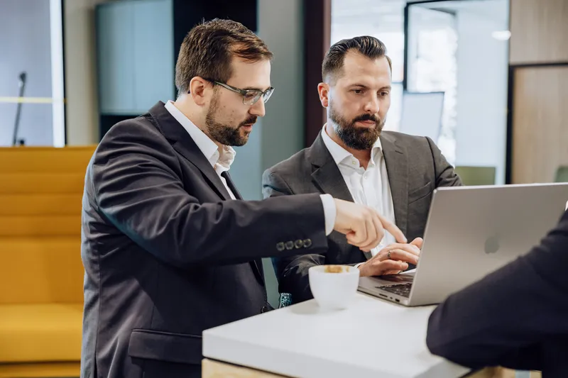 Two men in suits discussing something on a laptop at a table with a coffee cup.